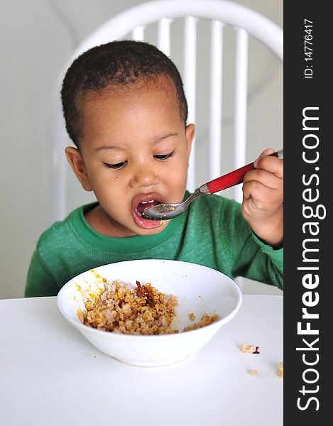 Young boy eating at table from white dish with red spoon. Young boy eating at table from white dish with red spoon