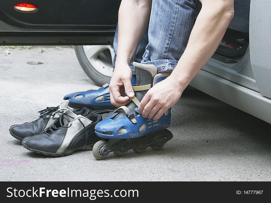 The man dresses the roller fads. Sits in the car. Near to rollers there is a footwear. In a frame of a hand, a foot and footwear.