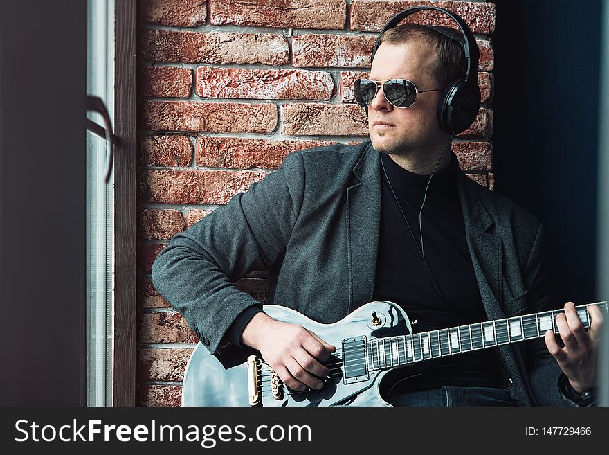 Man singer sitting on a window sill in a headphones with a guitar recording a track in a home studio. Man wearing sunglasses, jeans, black shirt and a jacket