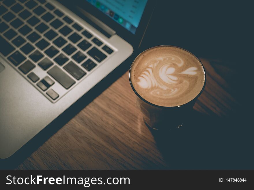 A cup of well-made latte with flower art on foam next to a laptop. A cup of well-made latte with flower art on foam next to a laptop