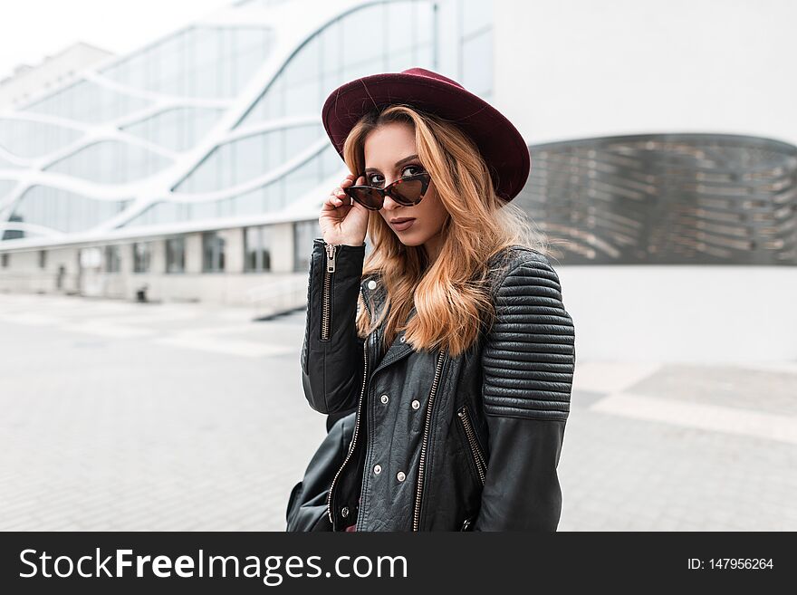 Sexy pretty young hipster woman in sunglasses in vintage hat in trendy leather jacket with black backpack stands