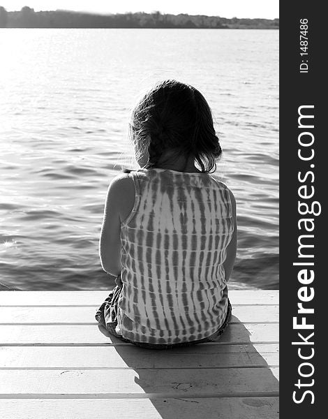Black and white image of a girl sitting on a pier, overlooking a lake. Black and white image of a girl sitting on a pier, overlooking a lake