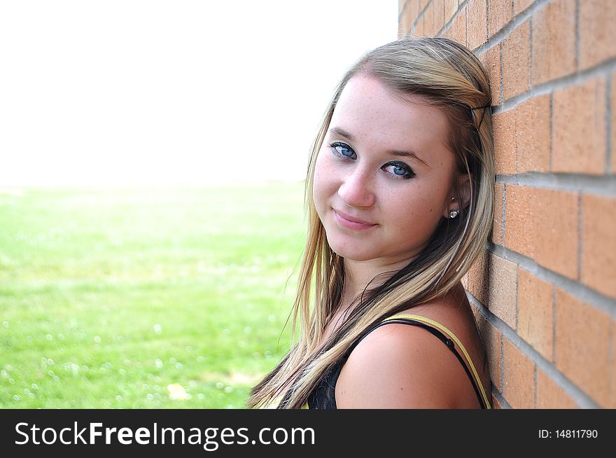 Happy young blond teenage girl standing in front of a brick wall smiling with white copyspace and green grass in the background behind her. Happy young blond teenage girl standing in front of a brick wall smiling with white copyspace and green grass in the background behind her.