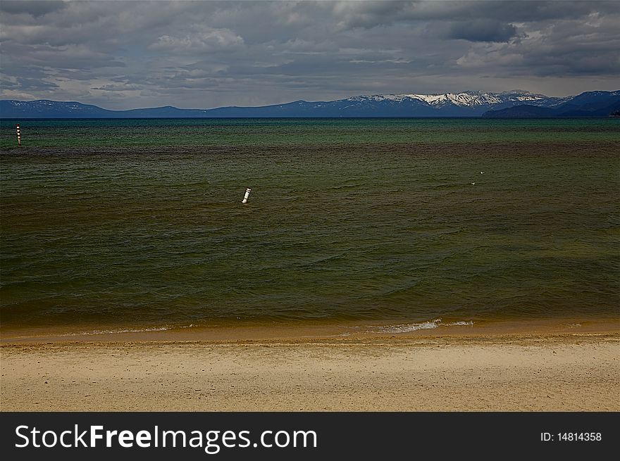 A sandy beach on Lake Tahoe Beach, Nevada.  The mountains are in the background and is a cloudy day.  The water has hues of green, brown and yellow.