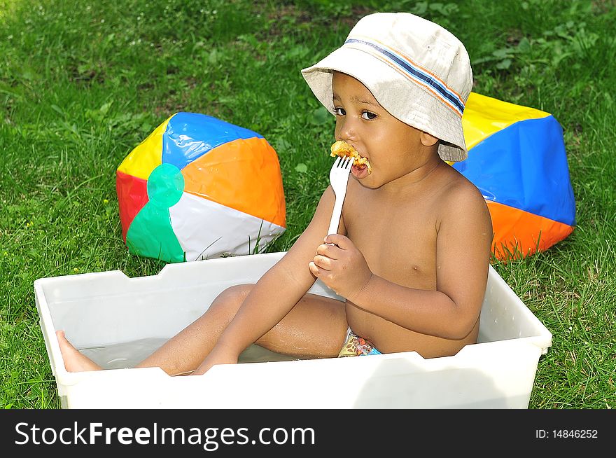 Young child eating while sitting in water with beech balls lying around him on grass. Young child eating while sitting in water with beech balls lying around him on grass