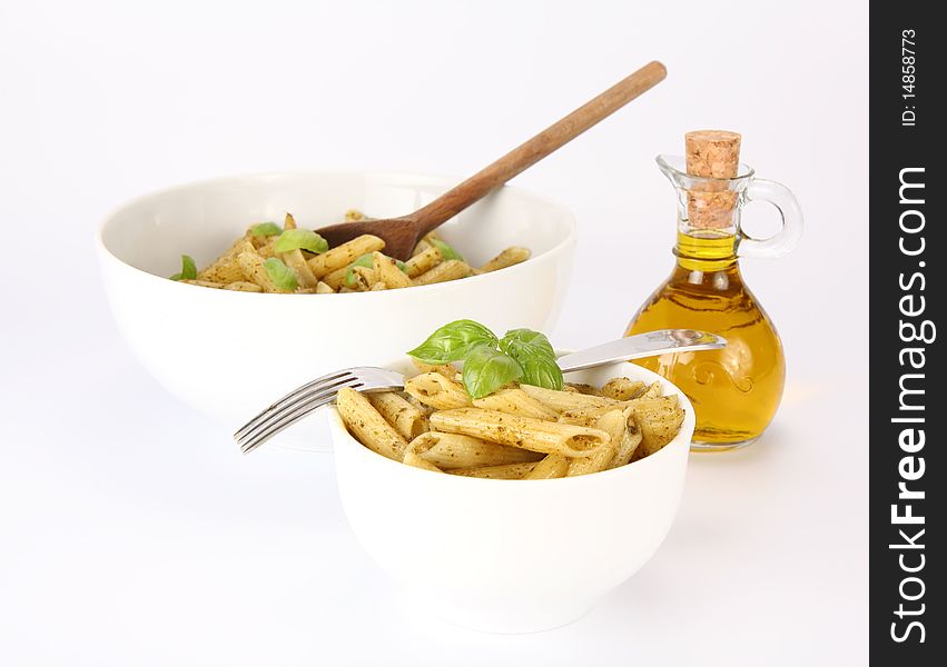 Penne with pesto decorated with basil and a fork in a small bowl and and a big bowl with wooden ladle , and a bottle of olive oil