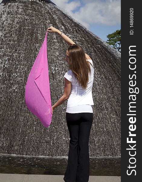 A woman standing with her back to the camera by a water fountain in the park while letting the wind blow her pink sarong. A woman standing with her back to the camera by a water fountain in the park while letting the wind blow her pink sarong.