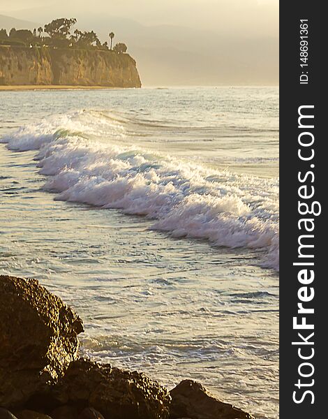 Foaming wave breaking onto ocean espanse with cliff and boulders at sunrise