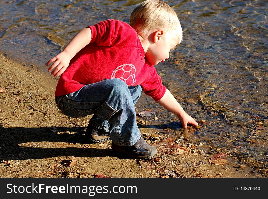 Young boy bends down to reach for a stone at the water's edge. Young boy bends down to reach for a stone at the water's edge.