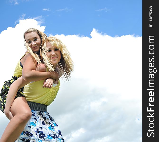 Two beautiful and attractive blond teenage girls giving a piggyback ride in the sunshine with clouds and sky in the background. Two beautiful and attractive blond teenage girls giving a piggyback ride in the sunshine with clouds and sky in the background.