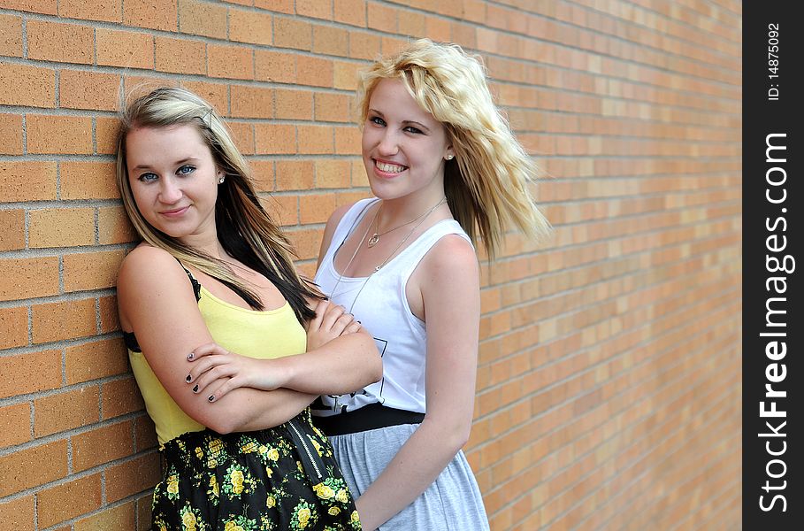 Happy young blond teenage girl friends standing in front of a brick wall smiling with each other. Happy young blond teenage girl friends standing in front of a brick wall smiling with each other.