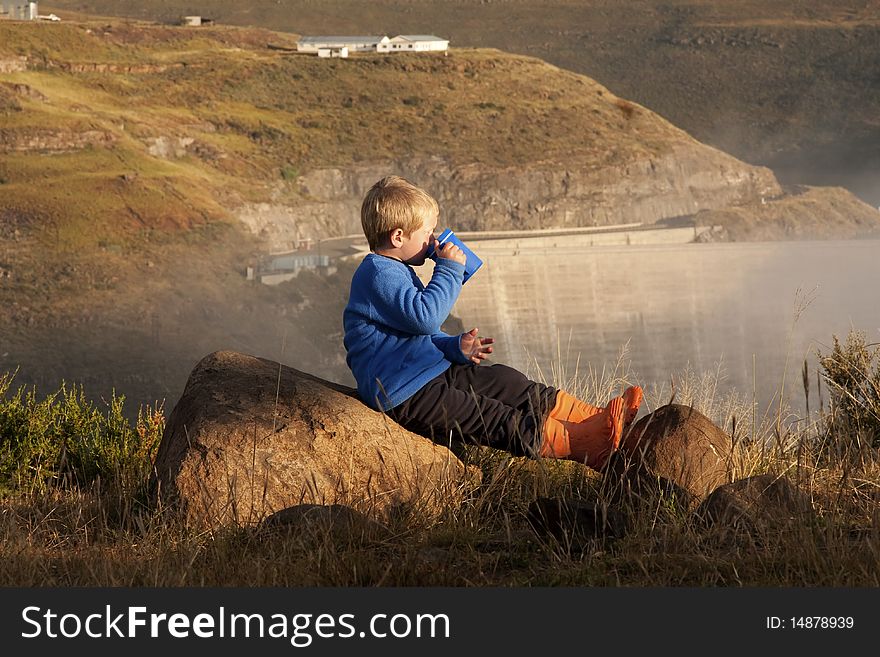 Young boy drinking hot drink in early morning light with Katse dam wall in Lesotho in background