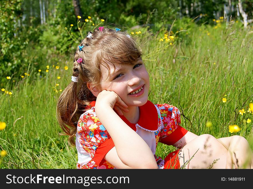 In the summer in park on a green grass with yellow flowers the smiling girl in a red dress sits. In the summer in park on a green grass with yellow flowers the smiling girl in a red dress sits