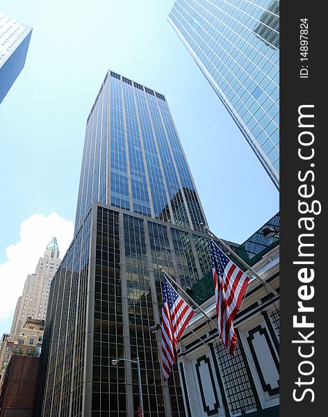 Glassy skyscrapers of Manhattan and american flags are photographed from below against blue sky. Glassy skyscrapers of Manhattan and american flags are photographed from below against blue sky.