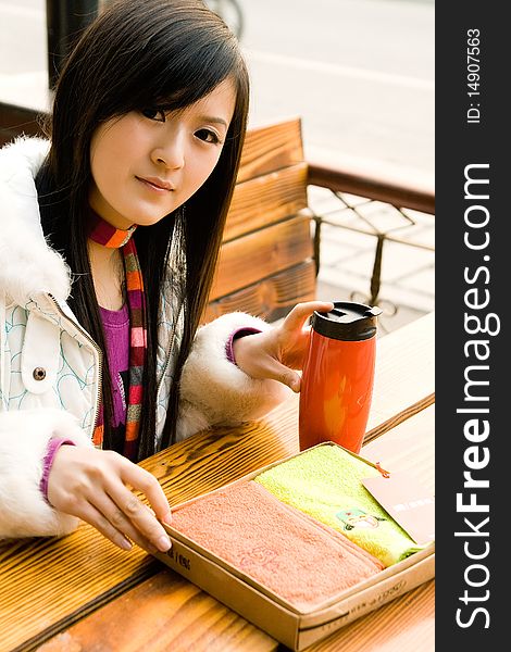 Beautiful girl sitting on bench in the street,in white coat,with a red cup and two new towels on the table