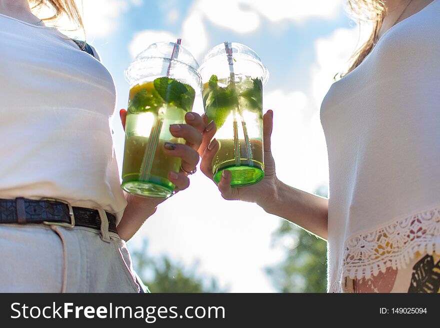 Two girls stand and hold plastic glasses with mojito, they clink glasses with drinks, close-up of refreshing drinks