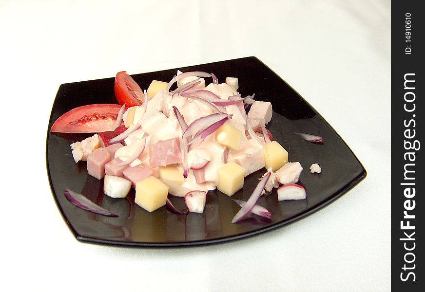 Salad with ham, cheese, redisos, chicken under cognac sauce, decorated with slices of tomato on a black square plate. Salad with ham, cheese, redisos, chicken under cognac sauce, decorated with slices of tomato on a black square plate