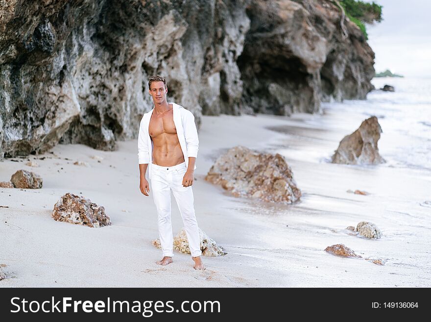 Sexy Tanned Man In White Shirt, Pants Rests On Beach, Ocean Waves At Background