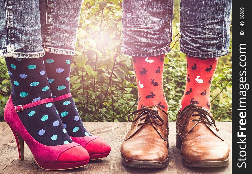 Men`s and women`s legs in fashionable shoes, bright, multi-colored socks on a wooden terrace on the background of green trees and sunny rays. Close-up. Concept of Style, Fashion and Beauty. Men`s and women`s legs in fashionable shoes, bright, multi-colored socks on a wooden terrace on the background of green trees and sunny rays. Close-up. Concept of Style, Fashion and Beauty