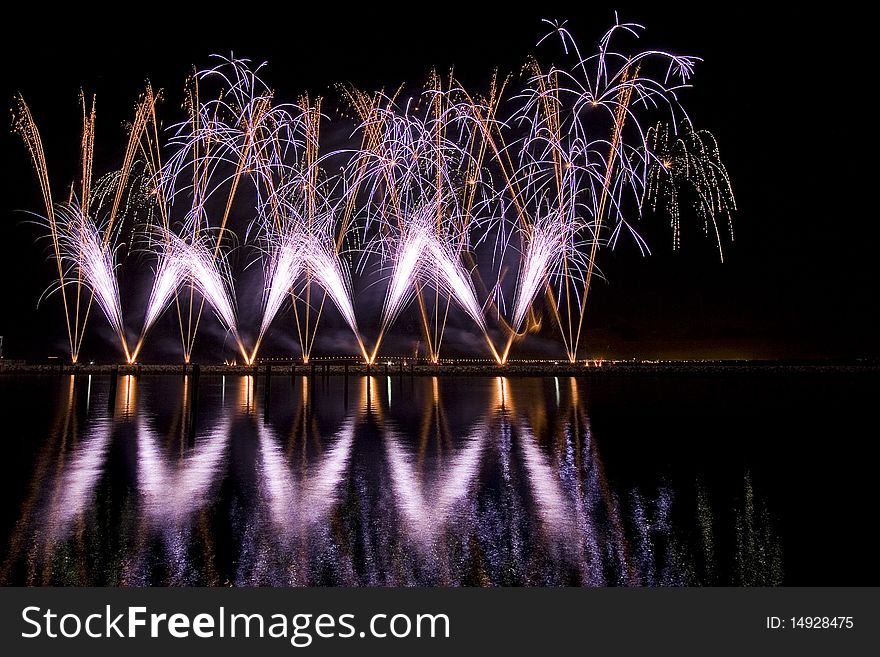 Long exposure photo of multiple fireworks over a lake, against a black sky.