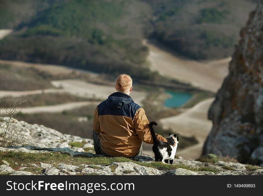 Redhead man in a brown-and-blue windbreaker sits on the slope of the Mangup plateau in the Crimea and strokes a black-and-white cat. Concept of travel, adventure, pets, animals and motivation. Redhead man in a brown-and-blue windbreaker sits on the slope of the Mangup plateau in the Crimea and strokes a black-and-white cat. Concept of travel, adventure, pets, animals and motivation.