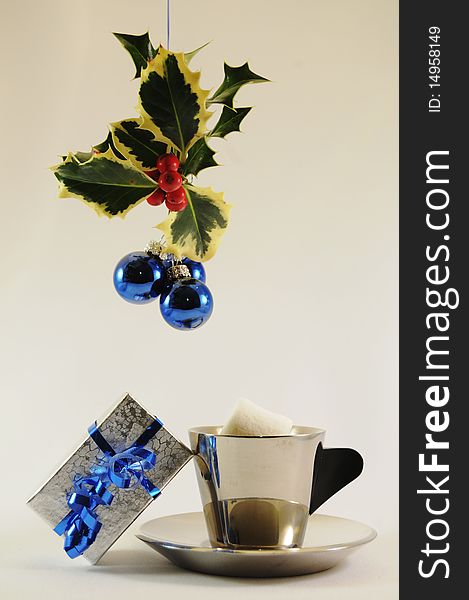Cup and saucer of cocoa with marshmellow with gift under holly with blue bulbs. Cup and saucer of cocoa with marshmellow with gift under holly with blue bulbs