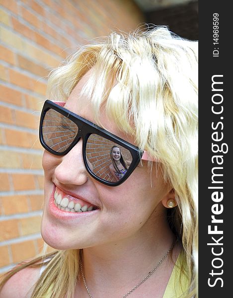 A girl smiling can be seen in her friend's sunglasses reflection against a brick wall. A girl smiling can be seen in her friend's sunglasses reflection against a brick wall.