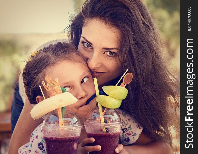 Happy kid girl and funny emotional mother drinking berries smoothie juice together in street summer outdoor cafe. Closeup toned portrait