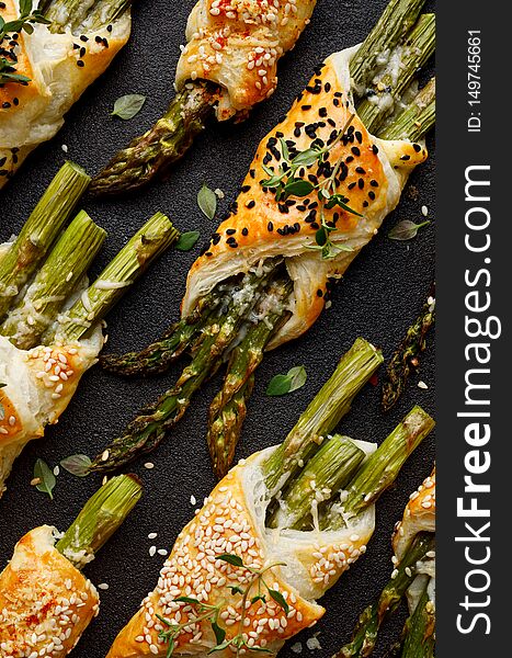 Baked green asparagus in puff pastry sprinkled with sesame seeds, nigella seeds and fresh thyme on a black background, close-up, t