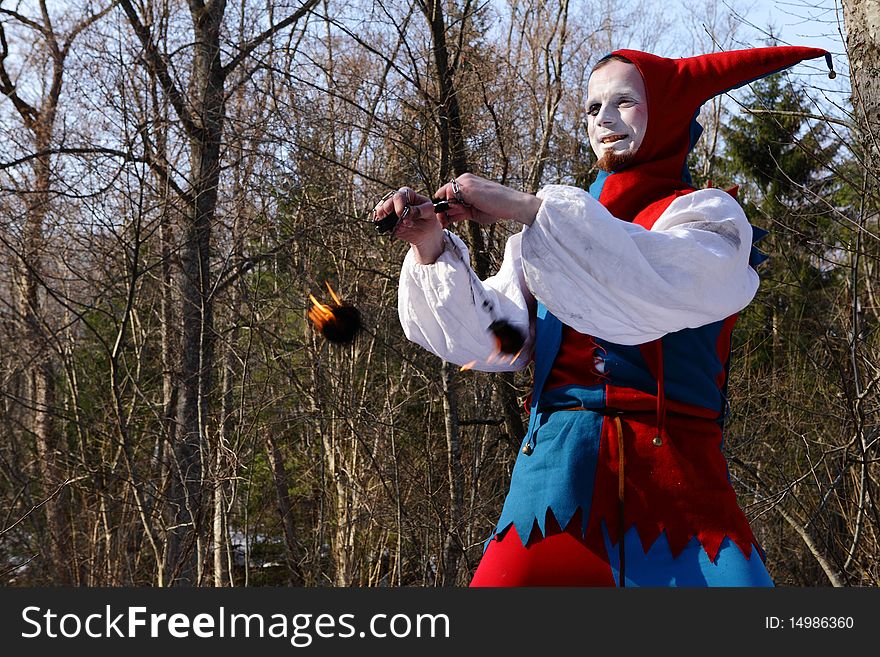 Artistic man in a suit of fire-clown rotate a fiery chain posing on backround of forest. Photo.