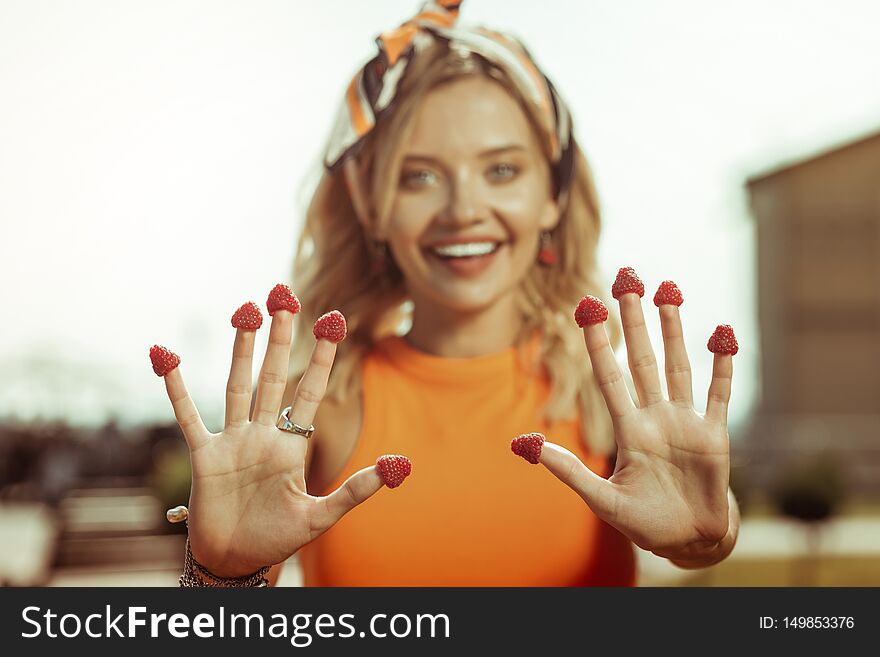 Strawberries on fingers. Close-up portrait of fair-haired smiling cheerful beautiful young woman wearing bright headband showing fingers with strawberries on them. Strawberries on fingers. Close-up portrait of fair-haired smiling cheerful beautiful young woman wearing bright headband showing fingers with strawberries on them.