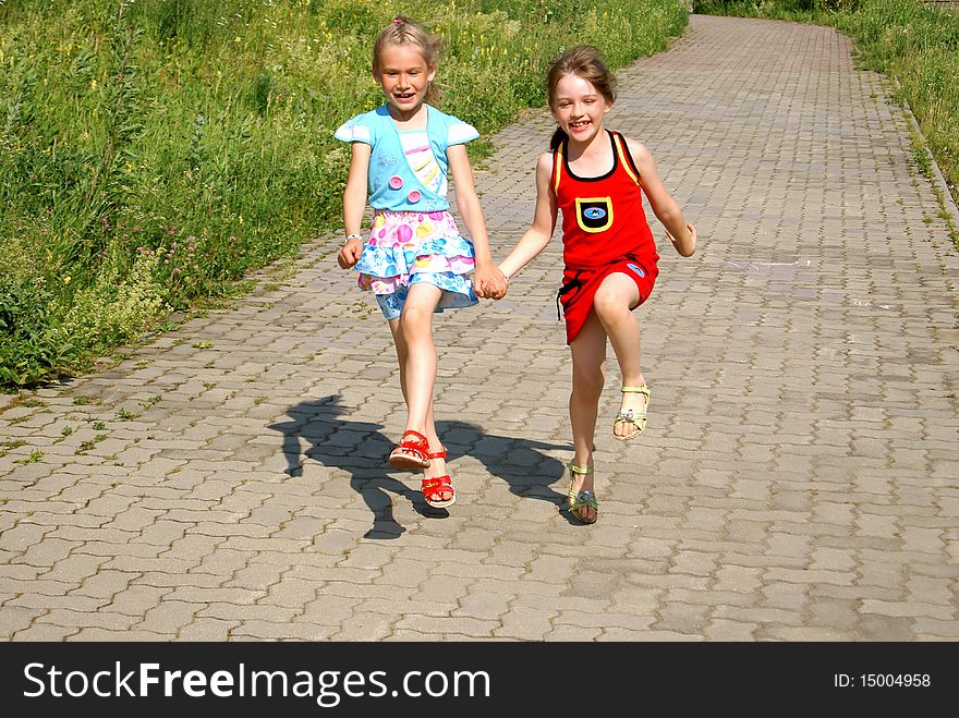 Two joyful girls keep for hands and run on sidewalk jumping up in the summer. Two joyful girls keep for hands and run on sidewalk jumping up in the summer