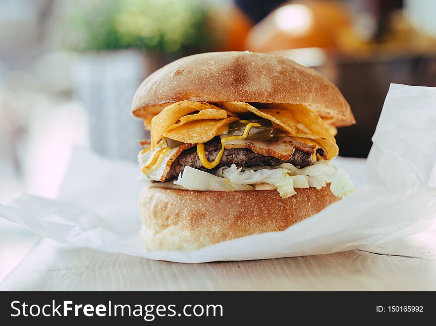 Fresh Close-up Of Designer Homemade Burger With Cutlet, Bacon, Chips And Mustard Sauce In Craft Paper On A Wooden Table