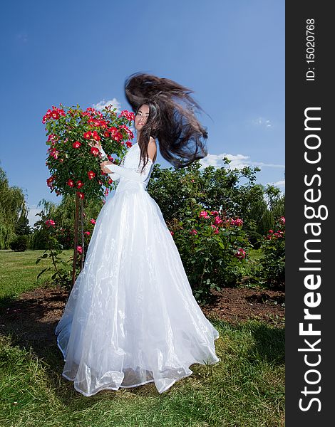 Romantic beautiful brunette bride standing alone in park near rosebush; hair waving