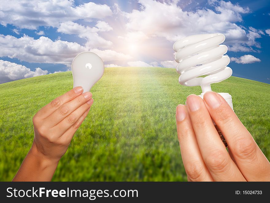 Female Hands Holding Energy Saving and Regular Light Bulbs Over Arched Horizon of Grass Field, Clouds and Sky. Female Hands Holding Energy Saving and Regular Light Bulbs Over Arched Horizon of Grass Field, Clouds and Sky.