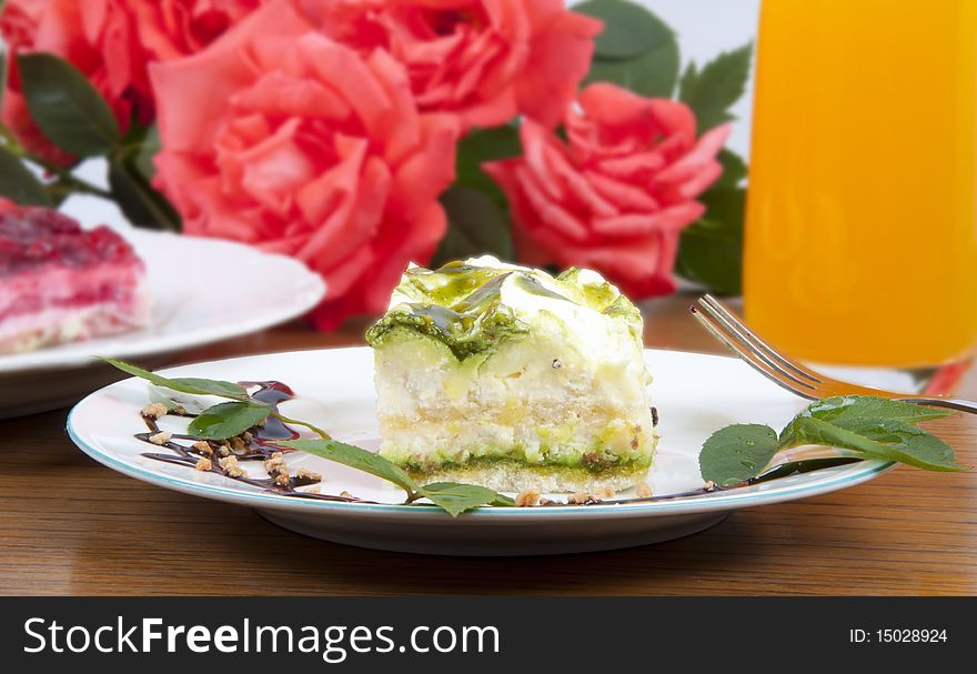 Sweet pistachio dessert with vanilla cream and nuts and chocolate on white plate with juice and rose on background.