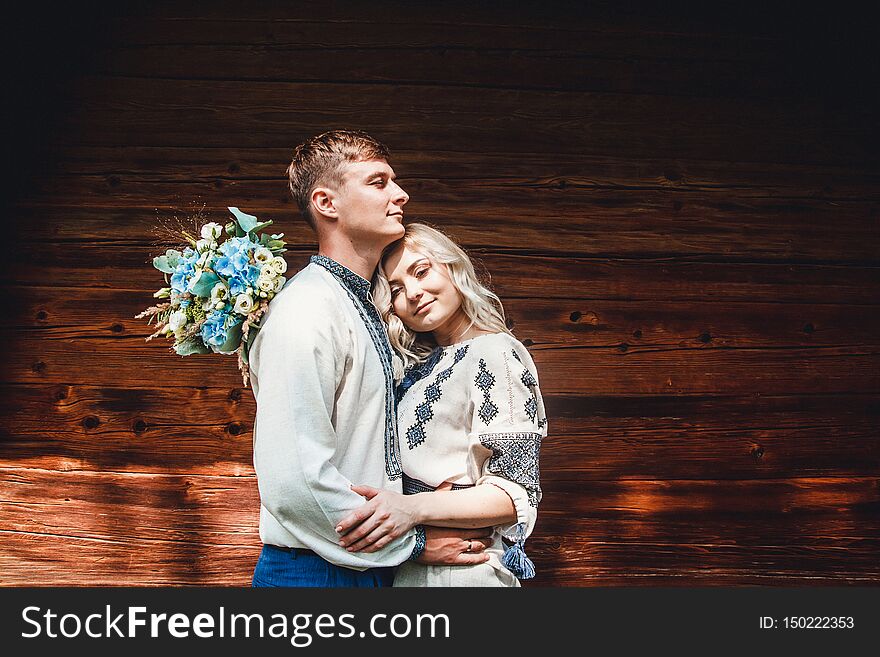 Amazing wedding couple in an embroidereds shirt with a bunch of flowers on the background of a wooden house.