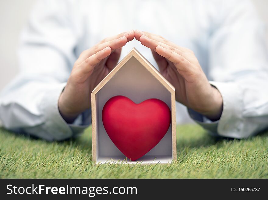 Wooden house with red heart on green grass protected by hands