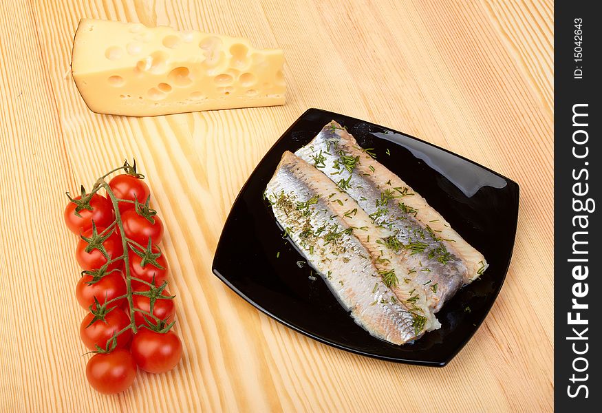 Herring on black plate with piece of cheese and tomato cherry on wood board