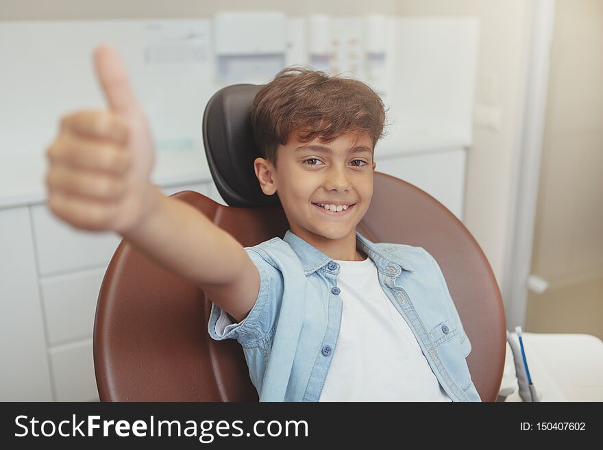 Lovely cheerful young boy smiling, showing thumbs up sitting in a dental chair. Adorable little boy looking happy after dental examination at the clinic. Lovely cheerful young boy smiling, showing thumbs up sitting in a dental chair. Adorable little boy looking happy after dental examination at the clinic