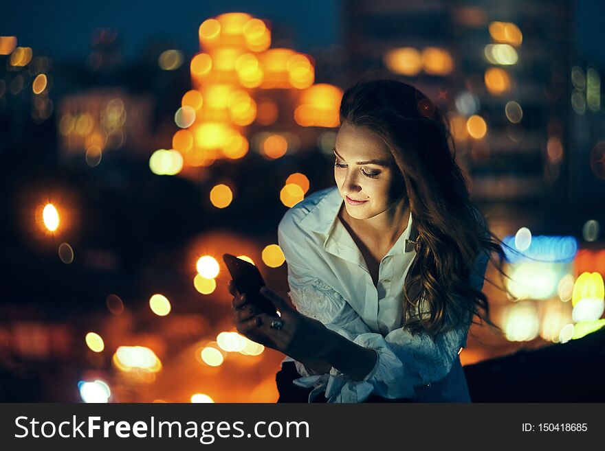 Young woman holding a smart phone in hands texting in the evening with city light on background. Young woman holding a smart phone in hands texting in the evening with city light on background