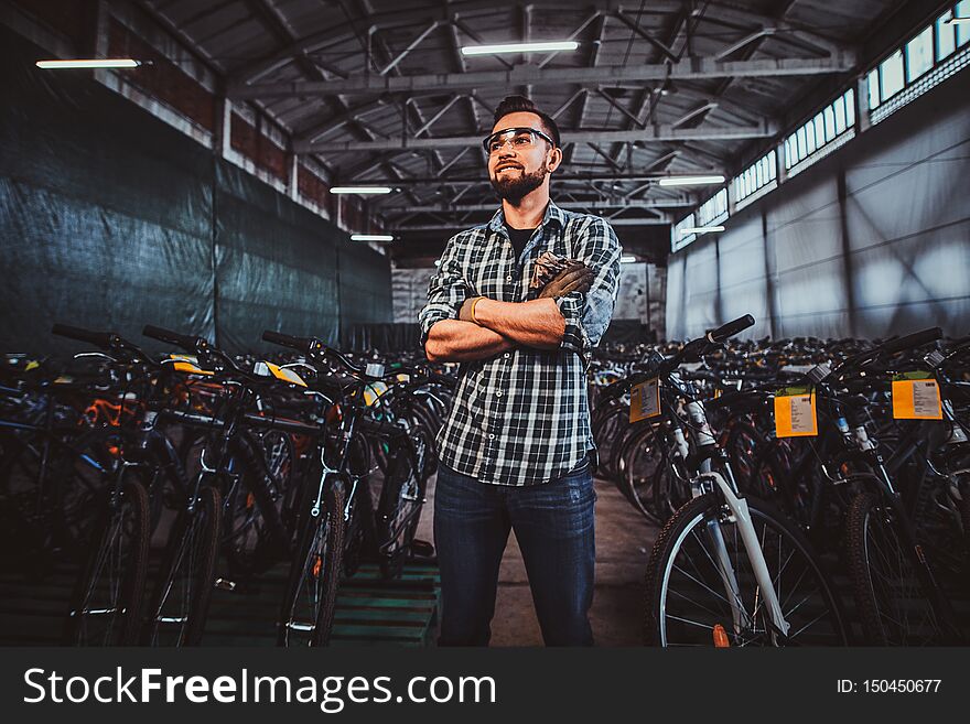 Pensive man in protective glasses and checkered shirt is posing at his own warehouse full of bicycles. Pensive man in protective glasses and checkered shirt is posing at his own warehouse full of bicycles.