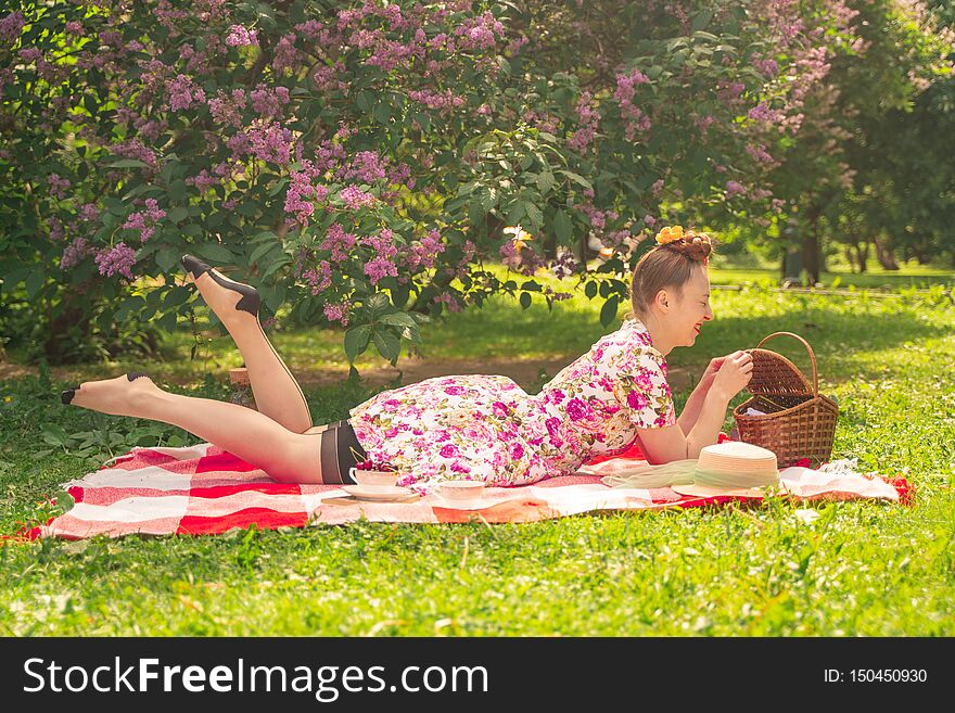 Sweetheart charming pinup girl in a summer dress on a checkered blanket in the Park near the bushes of lilac enjoys life and leisu
