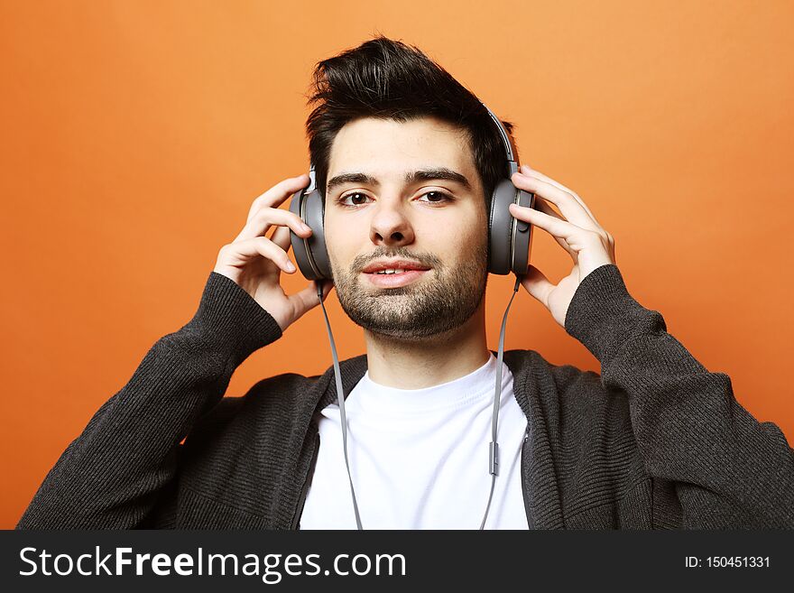 Happy young stylish man adjusting his headphones and smiling while standing against orange background