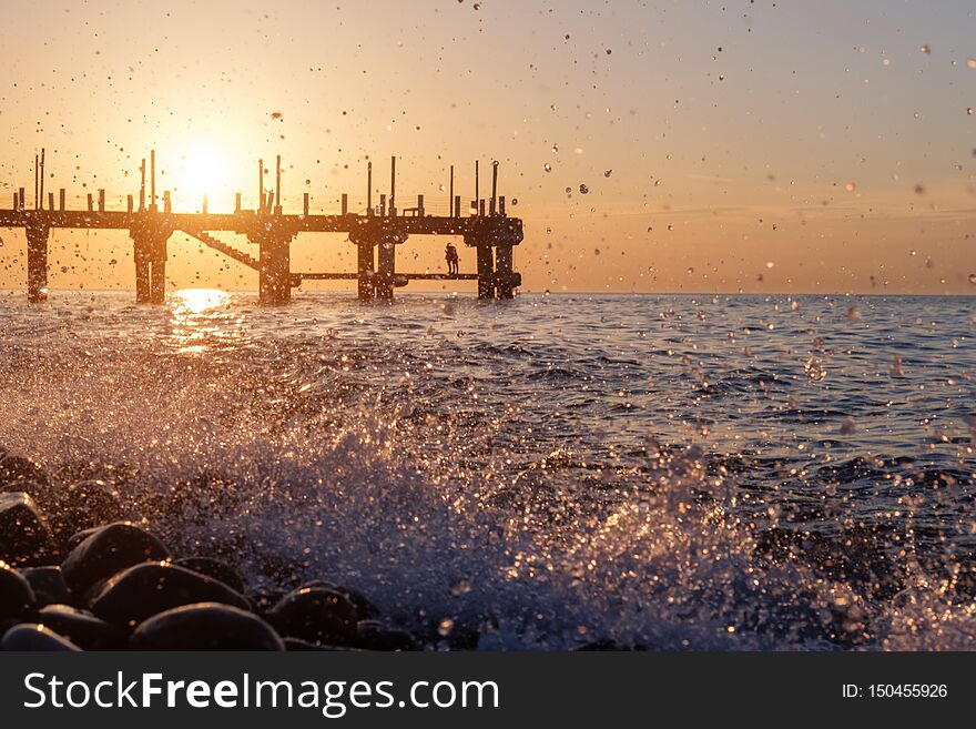 Splashing waves and blurry silhouette of couple on the pier at sunset