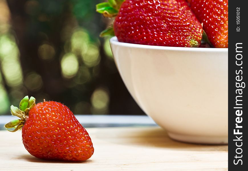 A single strawberry next to a white bowl of strawberries on a cutting board. A single strawberry next to a white bowl of strawberries on a cutting board