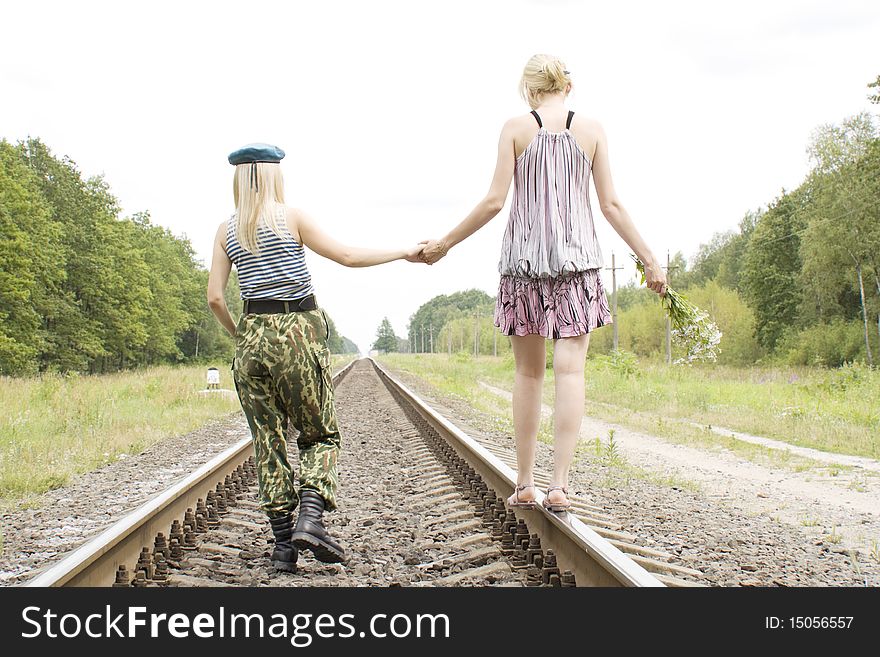 The woman in the military form goes on rails and holds a hand of the woman in a dress with a bouquet. The woman in the military form goes on rails and holds a hand of the woman in a dress with a bouquet