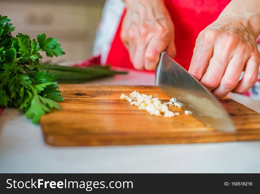 Woman`s hand with a knife slices garlic cloves on the wooden cutting board in the kitchen. Selective focus. Woman`s hand with a knife slices garlic cloves on the wooden cutting board in the kitchen. Selective focus