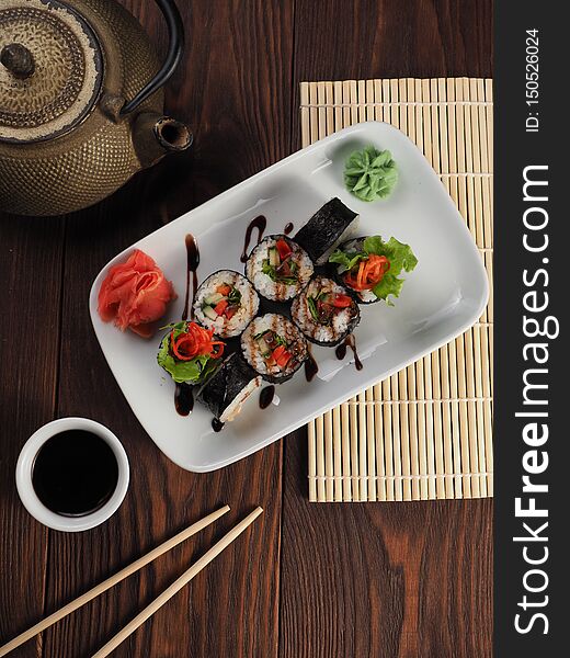 Fresh and tasty sushi on a white plate. Wooden table with a mat, chopsticks and kettle. Fresh and tasty sushi on a white plate. Wooden table with a mat, chopsticks and kettle
