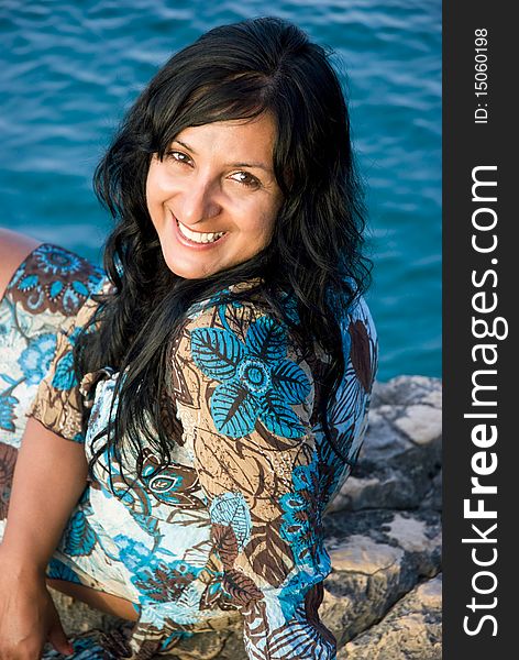 A beautiful happy smiling indian woman sitting on a rocky beach in Paxos Greece.